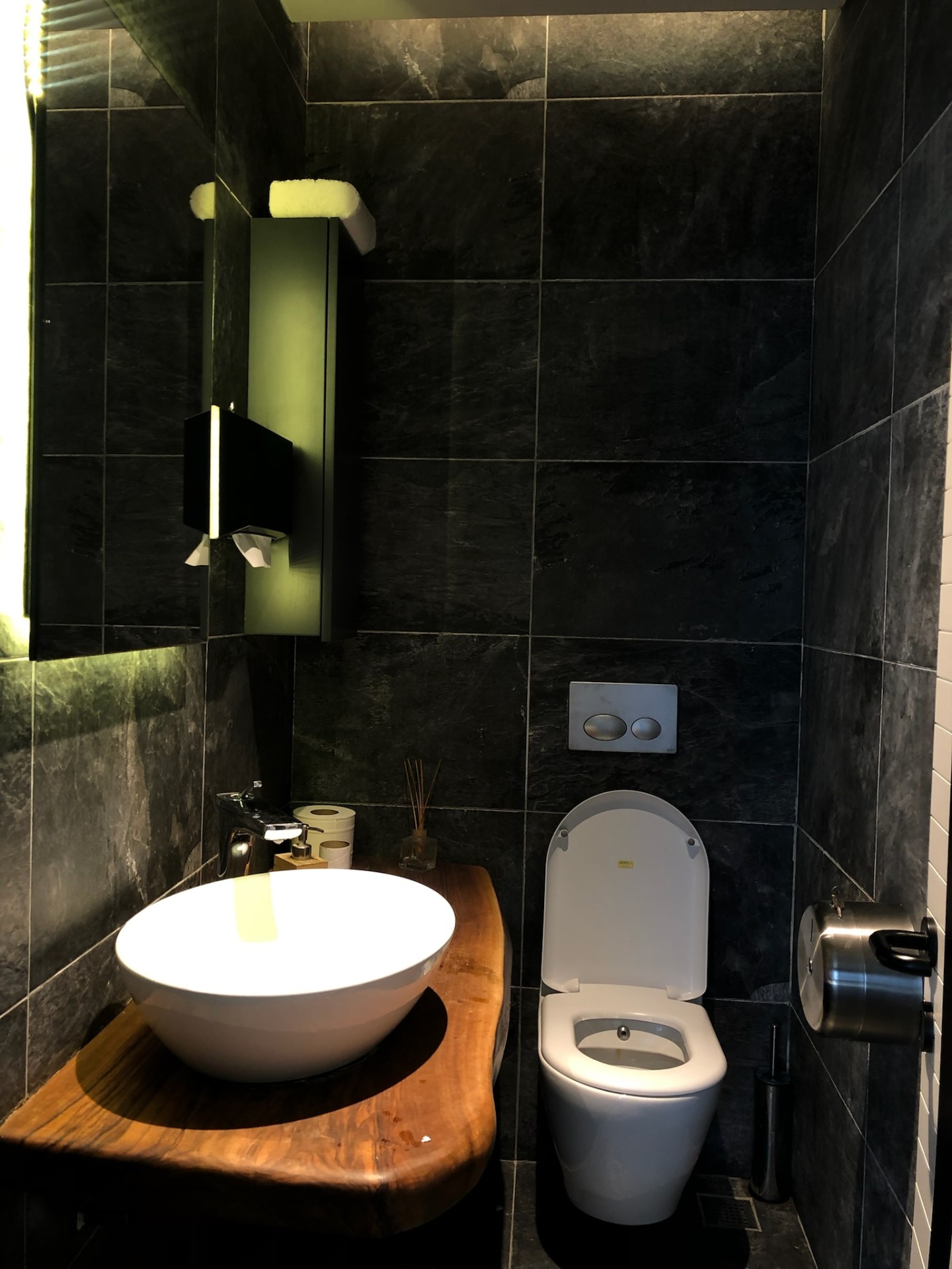 Restaurant WC in black slate with live-edge walnut counter and backlit vertical mirror.